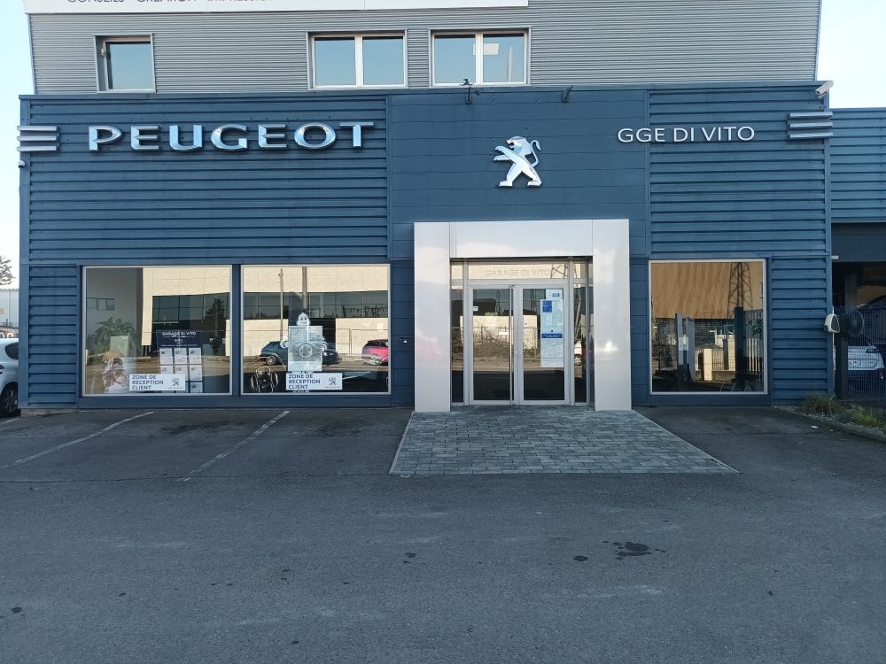 Extérieur d'une concession automobile Peugeot, bâtiment bleu et gris, logo au-dessus de l'entrée.