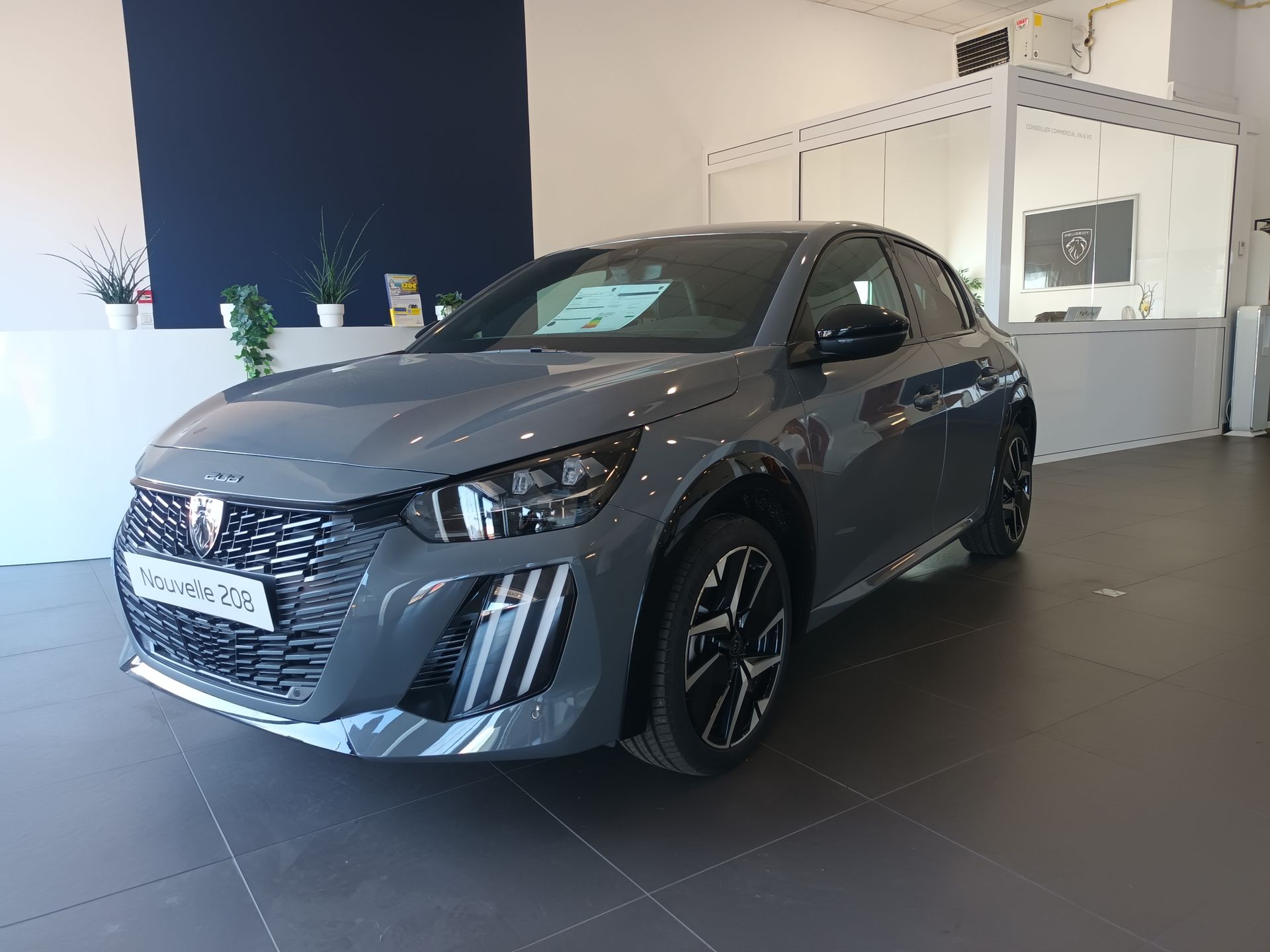 Peugeot 208 grise dans une salle d'exposition avec un fond blanc et bleu.
