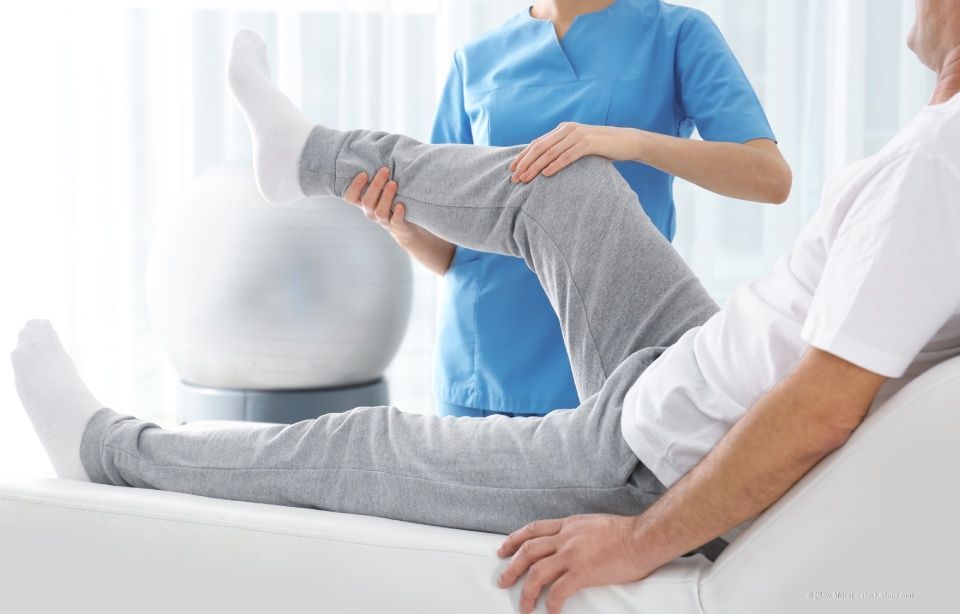 Physiotherapeut dehnt das Bein eines Patienten auf einer Behandlungsliege in einer hellen Klinik.