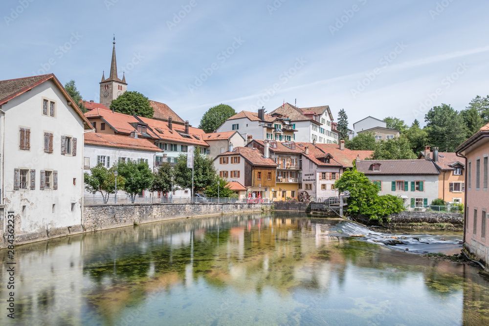 Une rivière s'écoule par-dessus un petit barrage de pierre vers un village niché au pied d'une montagne.