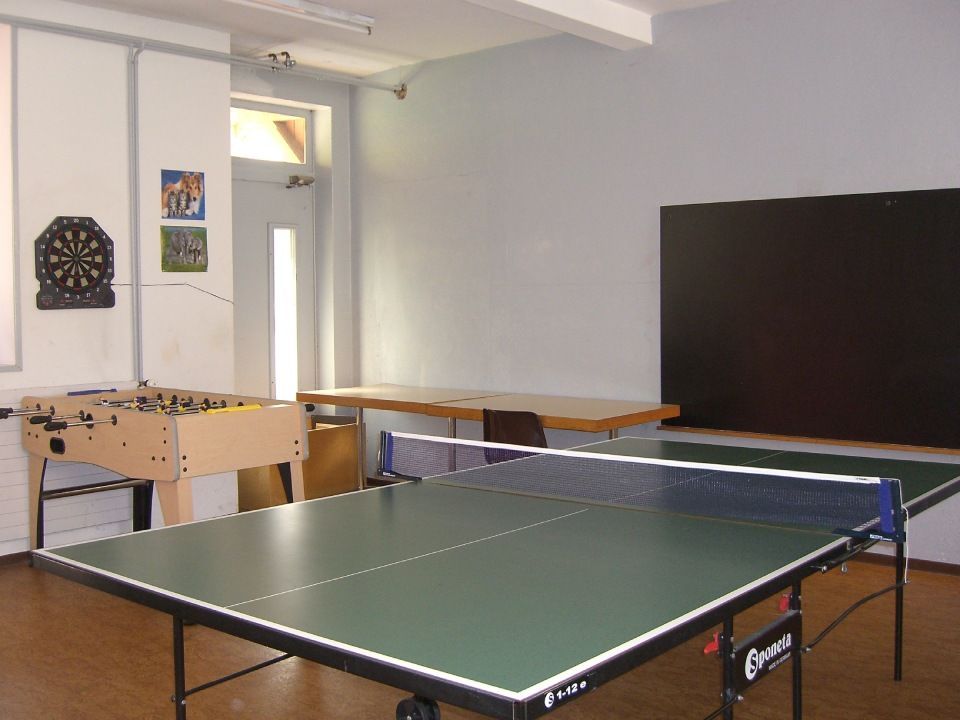 Une pièce avec une table de ping-pong verte, un baby-foot, un jeu de fléchettes et un long bureau en bois adossé à un mur nu.