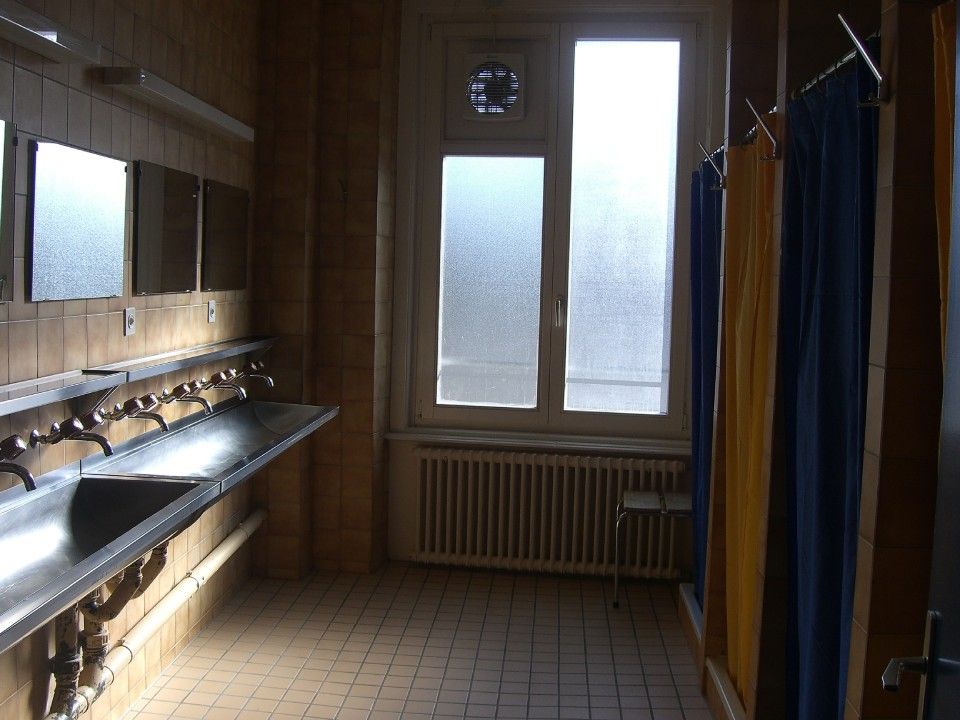 Des toilettes publiques avec un long lavabo en métal, des miroirs et des cabines de douche avec des rideaux jaunes et bleus.