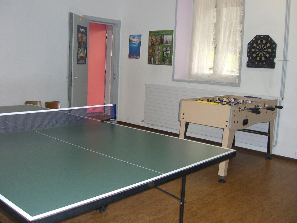 Une salle de jeux avec une table de ping-pong verte au premier plan, un baby-foot et une cible de fléchettes au mur.