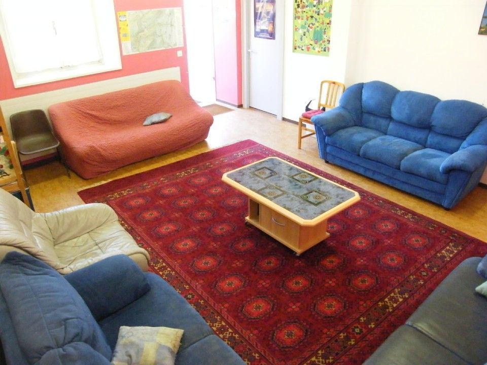 Un salon avec un tapis rouge, une table basse, un canapé rouge, un canapé bleu et un fauteuil couleur crème.
