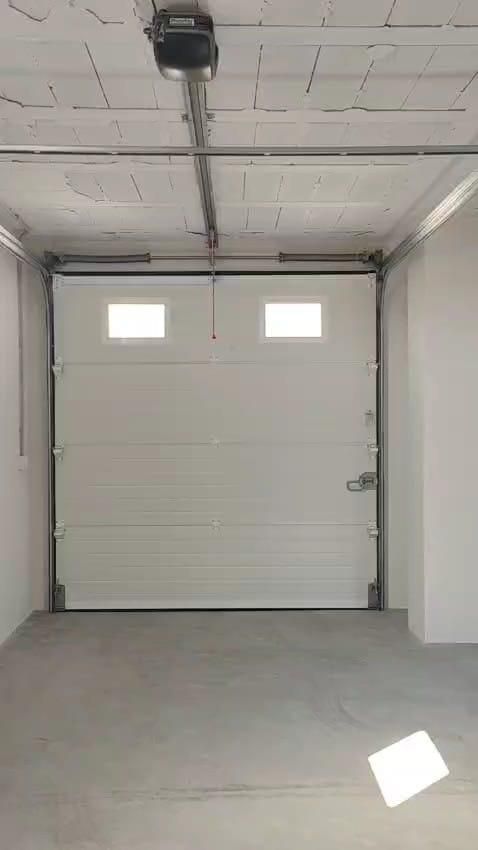 Puerta de garaje blanca, dos ventanas pequeñas, abiertas en un espacio con piso de concreto, abridor superior visible.