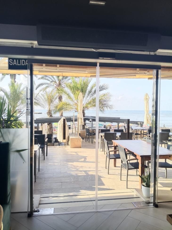 Vista del restaurante: Puertas de cristal transparente que dan a una playa con mesas, sillas y palmeras. El mar de fondo.