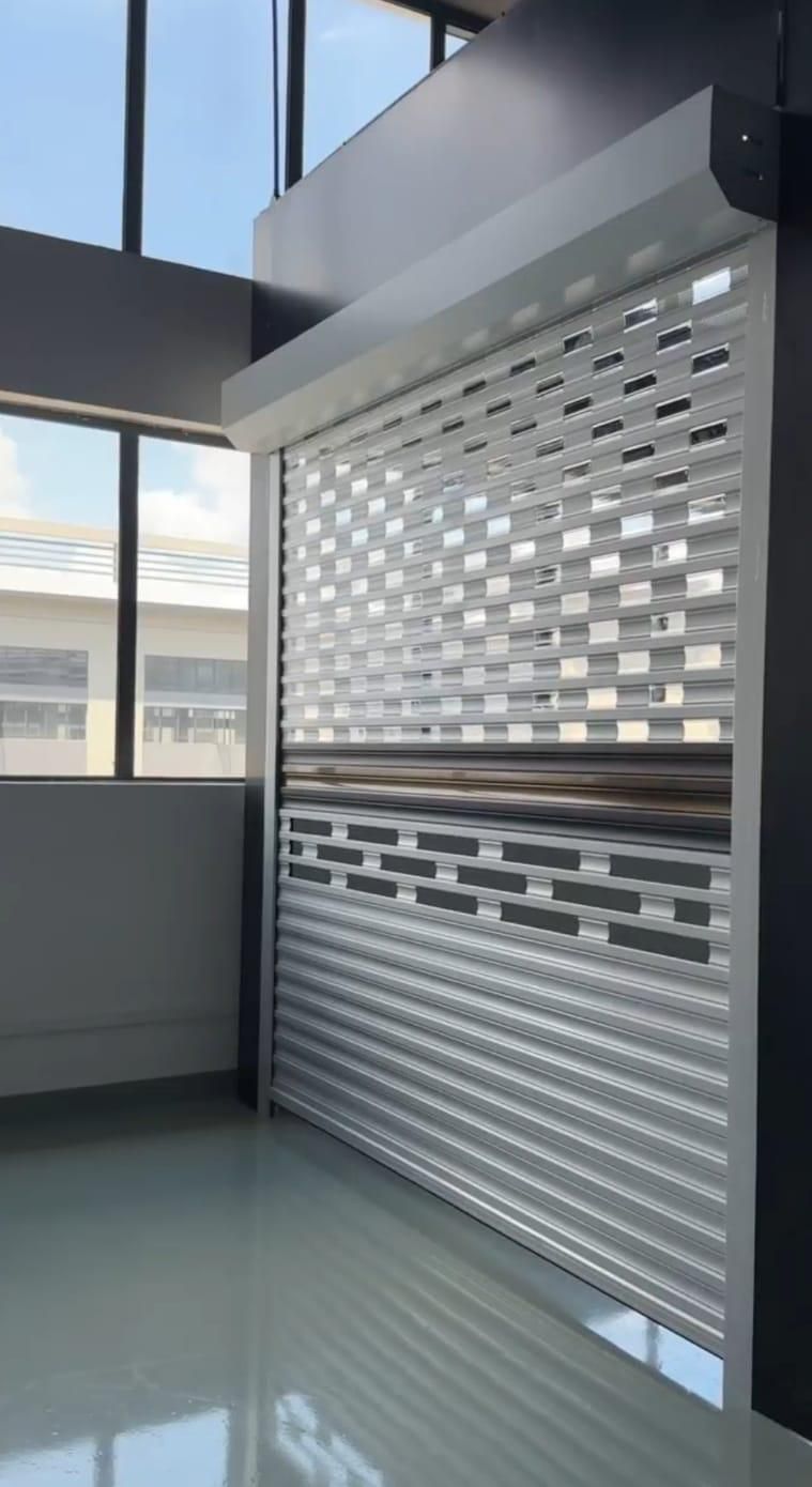 Persiana enrollable perforada de color blanco en nave industrial, con ventana visible en el lado izquierdo.
