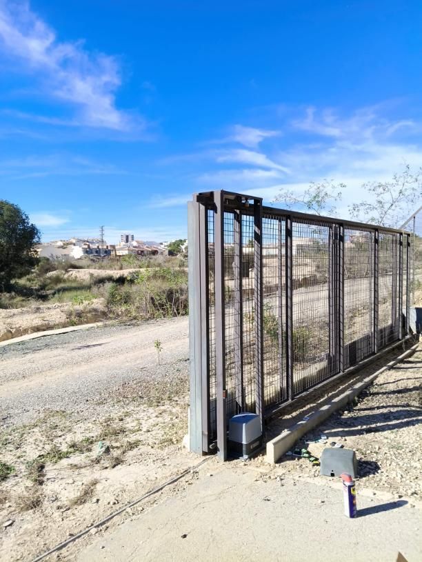 Puerta corrediza de metal sobre un camino de tierra con un pueblo distante bajo un cielo azul.
