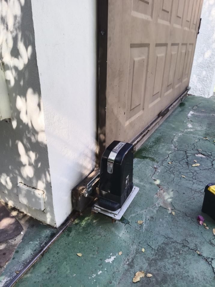 Abridor automático de puerta corredera sobre superficie de hormigón, junto a puerta de madera beige y pared blanca.