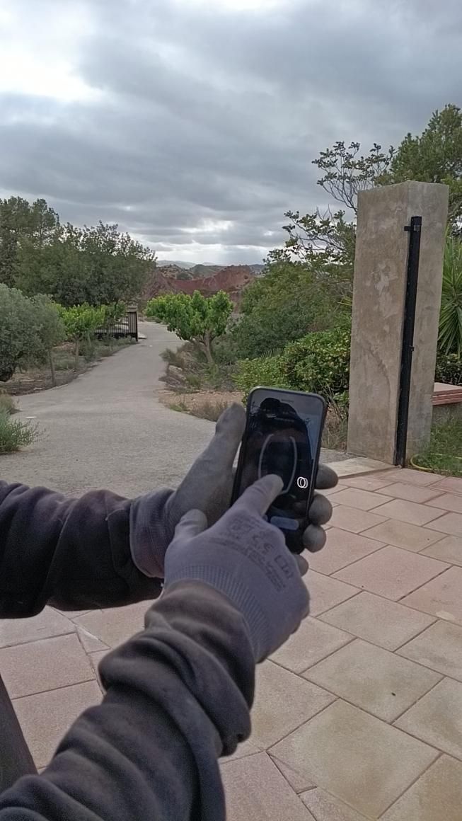 Persona con manos enguantadas que usa un teléfono para fotografiar un pilar de piedra y un camino
al aire libre.