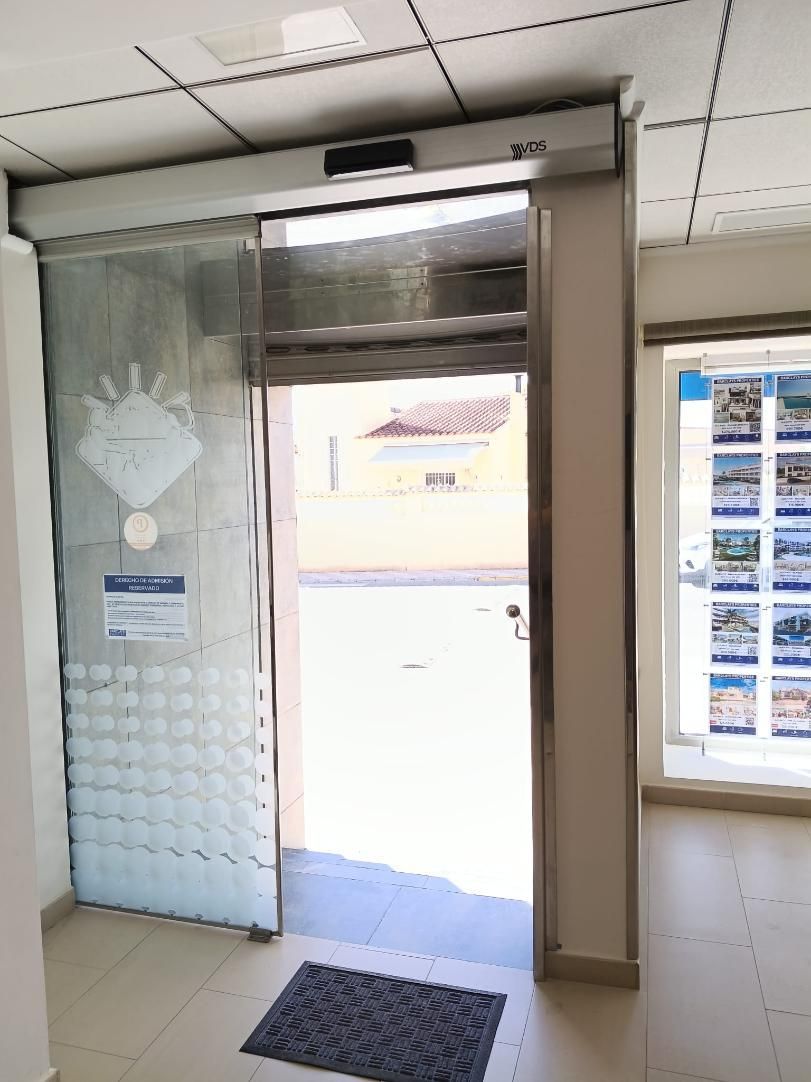 Puerta corrediza automática de vidrio que da al exterior, con alfombra en el piso y una ventana visible.