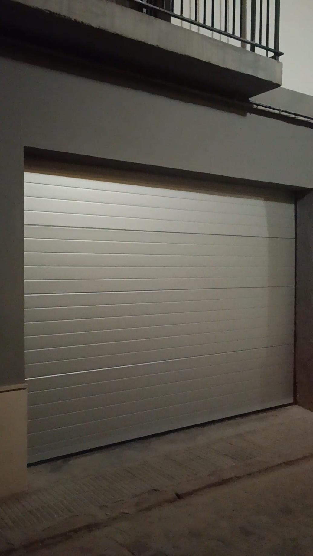 Puerta de garaje plateada con estrías horizontales. Exterior oscuro, suelo de hormigón.