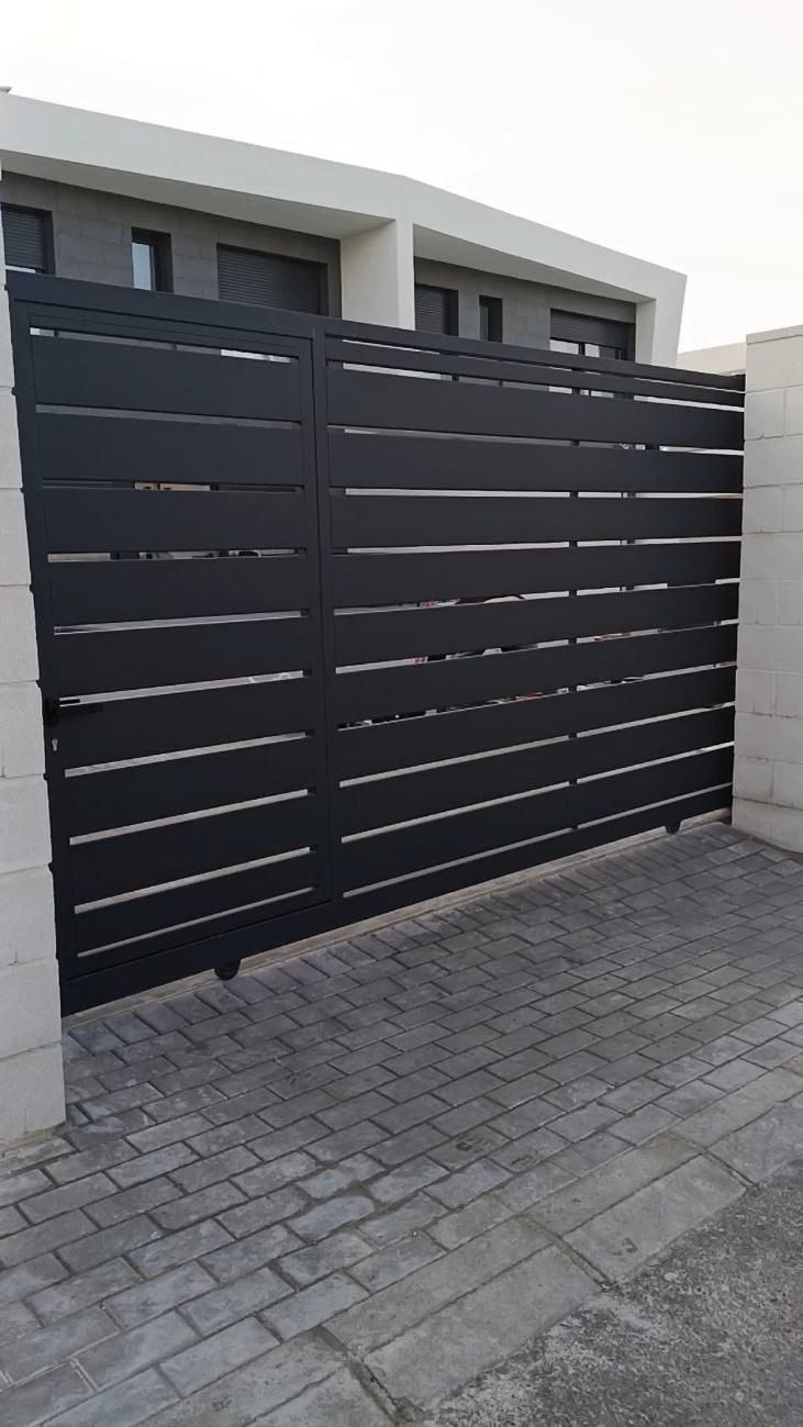 Puerta de lamas horizontales de color negro entre dos pilares de hormigón sobre adoquines de ladrillo.