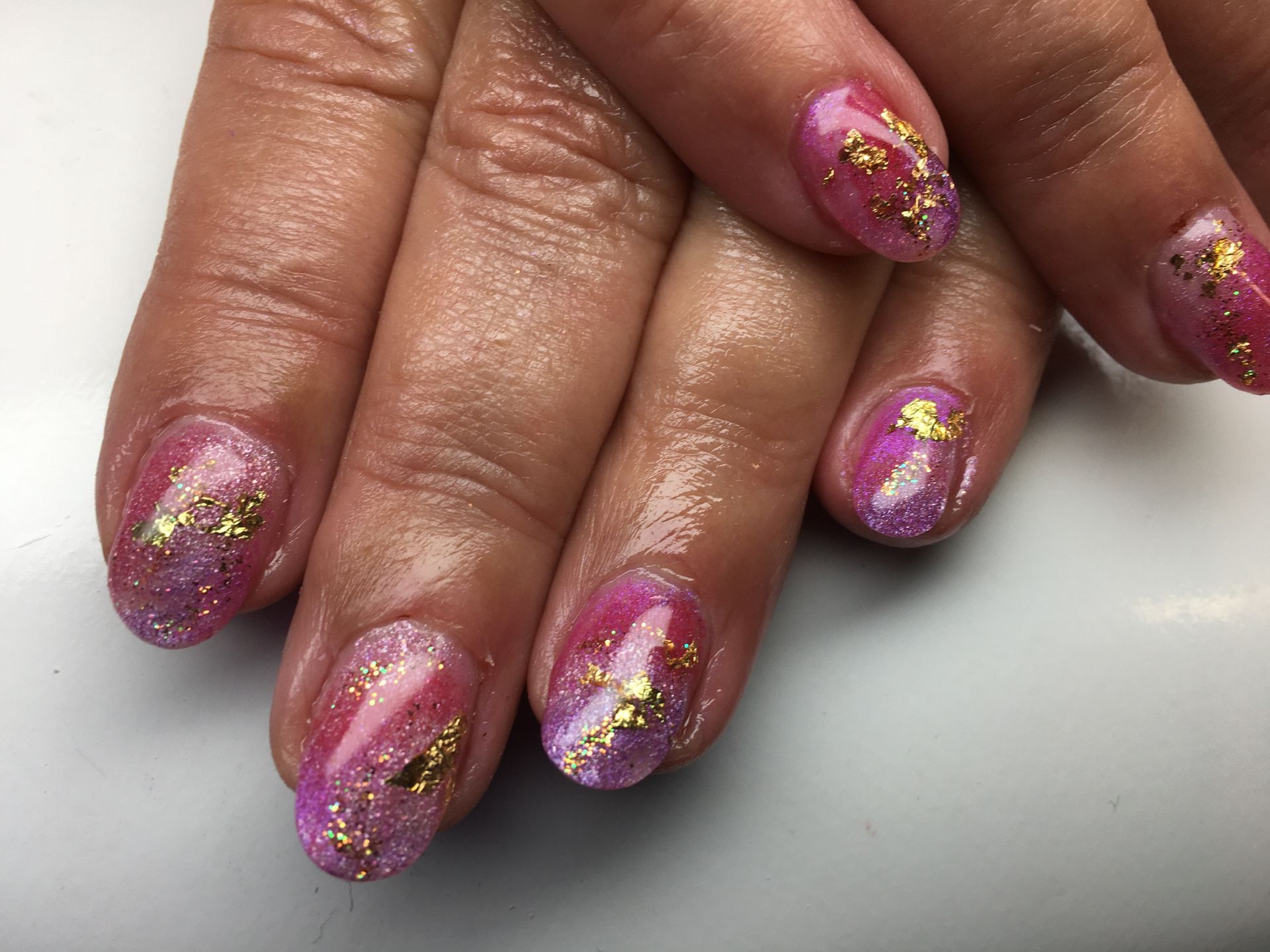 Pink-lila Glitzer Maniküre mit goldenen Folienakzenten auf kurzen, rund gefeilten Nägeln.