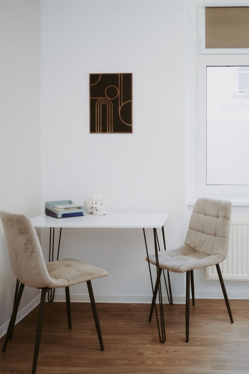 Kleiner weißer Tisch mit zwei beigen Stühlen vor einer weißen Wand mit einem gerahmten abstrakten Kunstwerk. Fenster rechts.