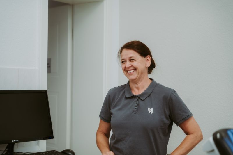 Eine Frau in einem grauen Poloshirt lächelt in einem weiß getünchten Raum.