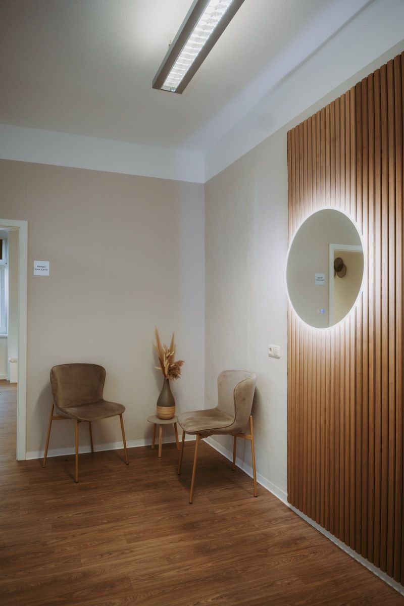 Wartebereich mit Stühlen, dekorativer Wand mit beleuchtetem runden Spiegel und Holzboden.