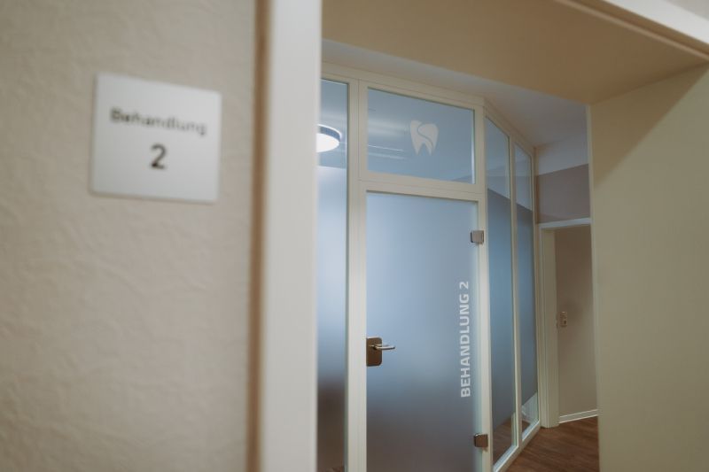 Schild mit der Aufschrift „Behandlung 2“ neben einer Milchglastür mit dem Logo einer Zahnklinik.