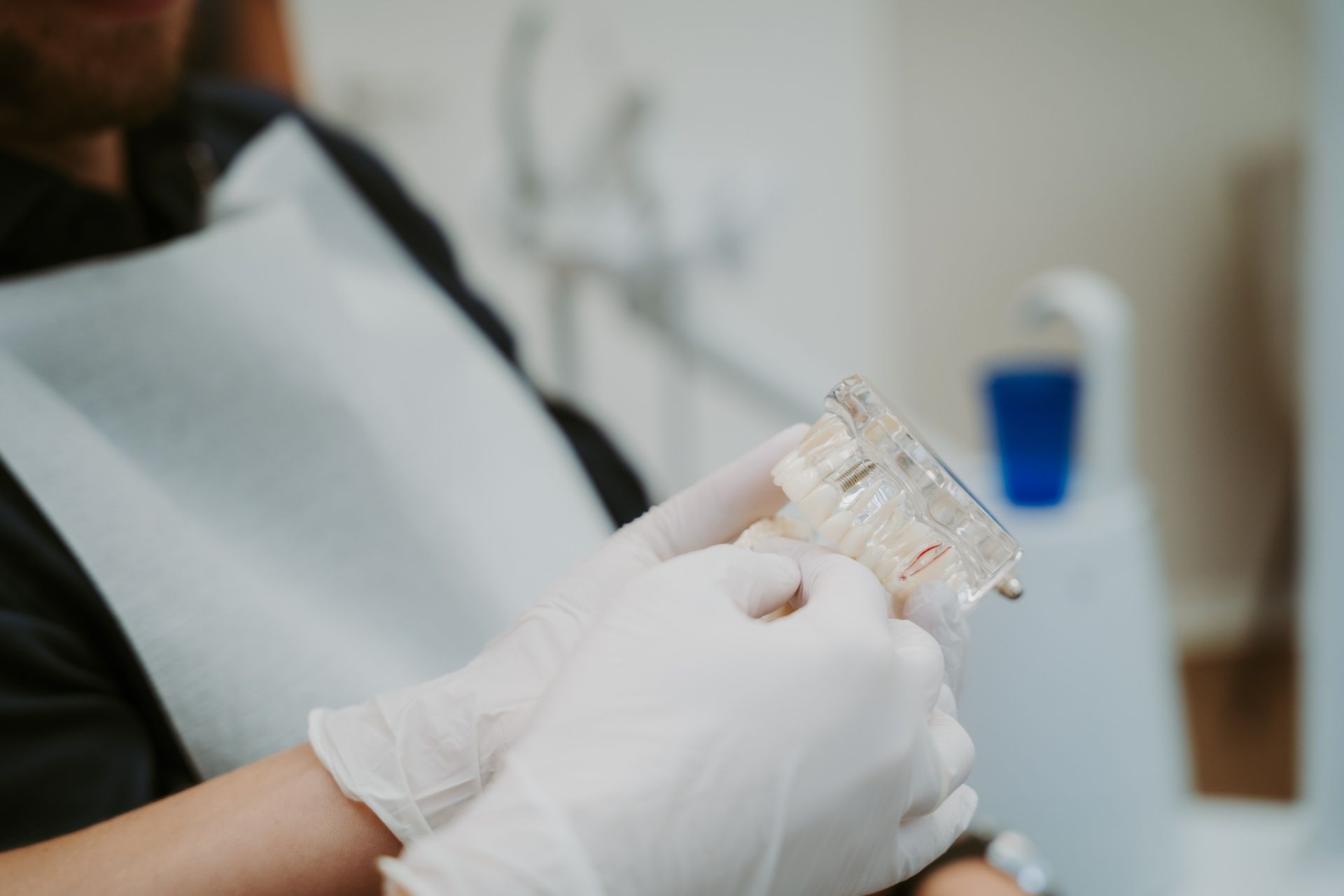 Ein Zahnarzt mit Handschuhen hält ein transparentes Zahnmodell. Im Hintergrund ist ein Patient mit Lätzchen
