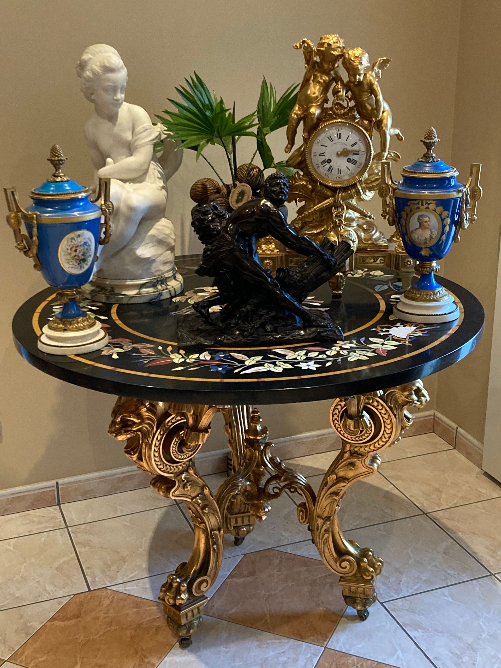 Table ronde ornée avec pieds dorés, plateau noir décoré de figurines, horloge et vases bleus.