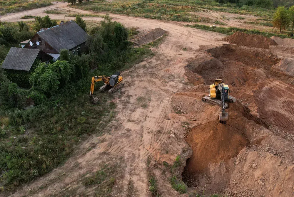 Dos excavadoras trabajando en un campo de tierra cerca de una casa con techo desgastado.