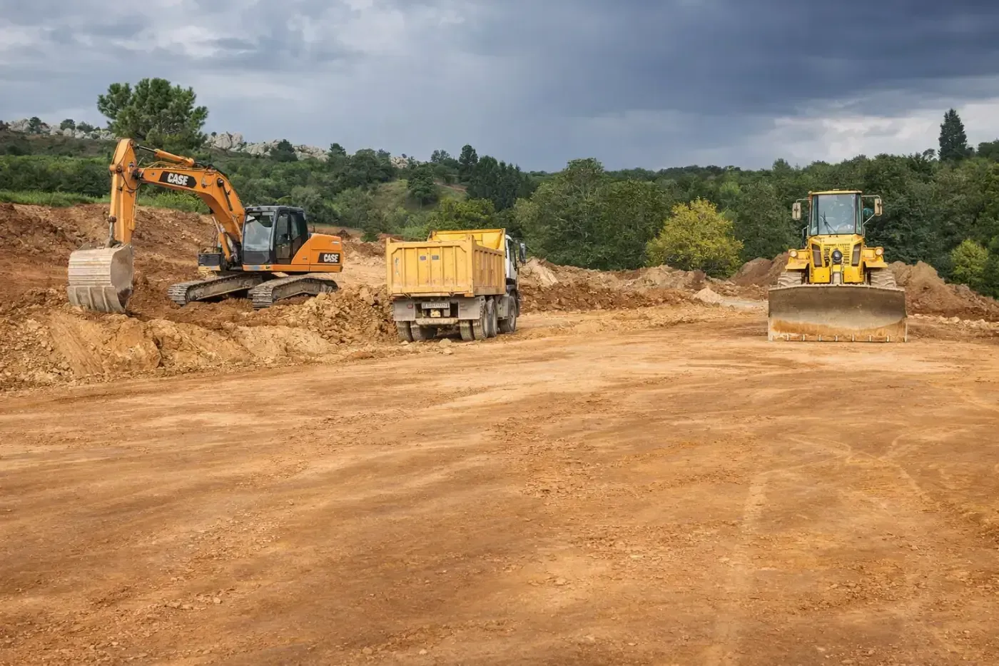 Obra con excavadora y bulldozer moviendo tierra, con camión volquete entre ambos. Cielo nublado.