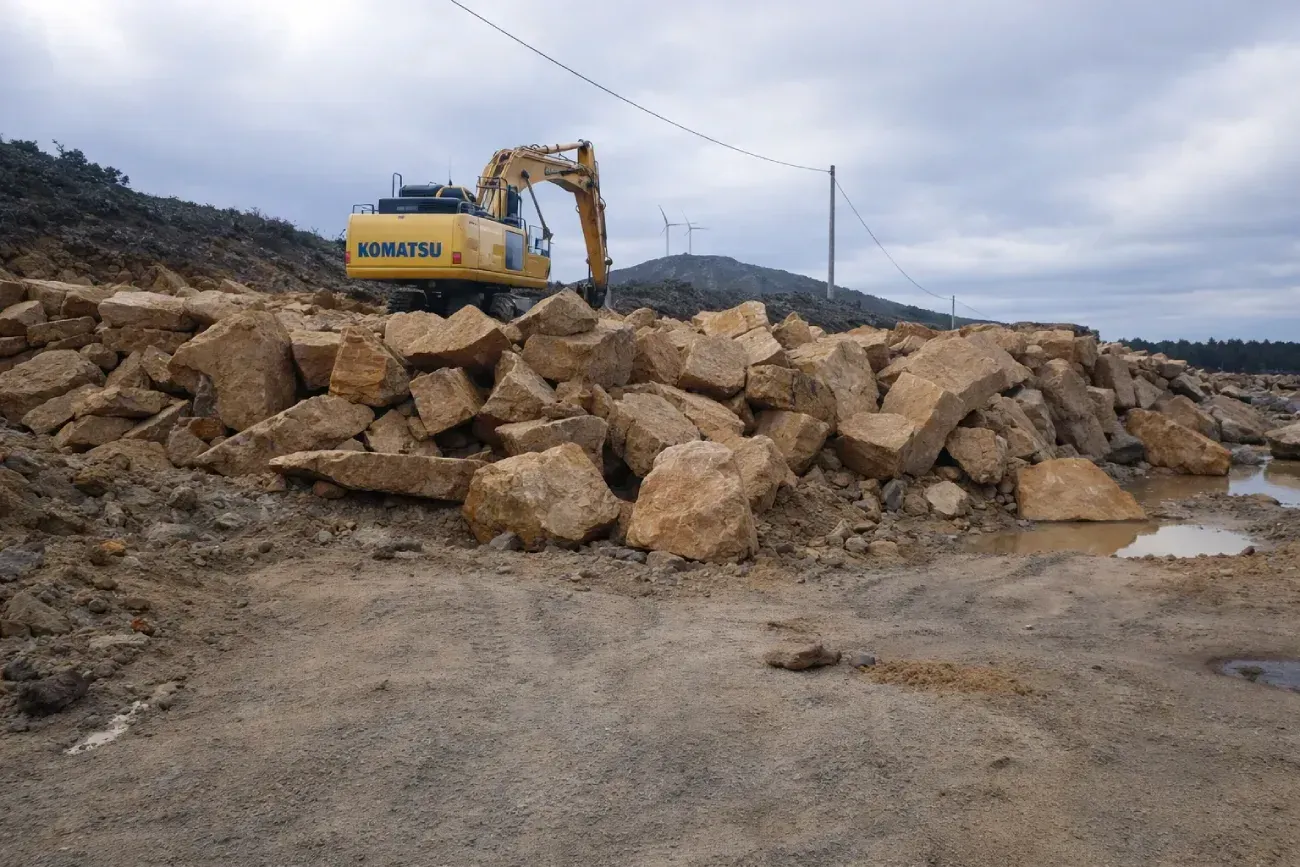 Dos excavadoras trabajando en un campo de tierra cerca de una casa con techo desgastado.