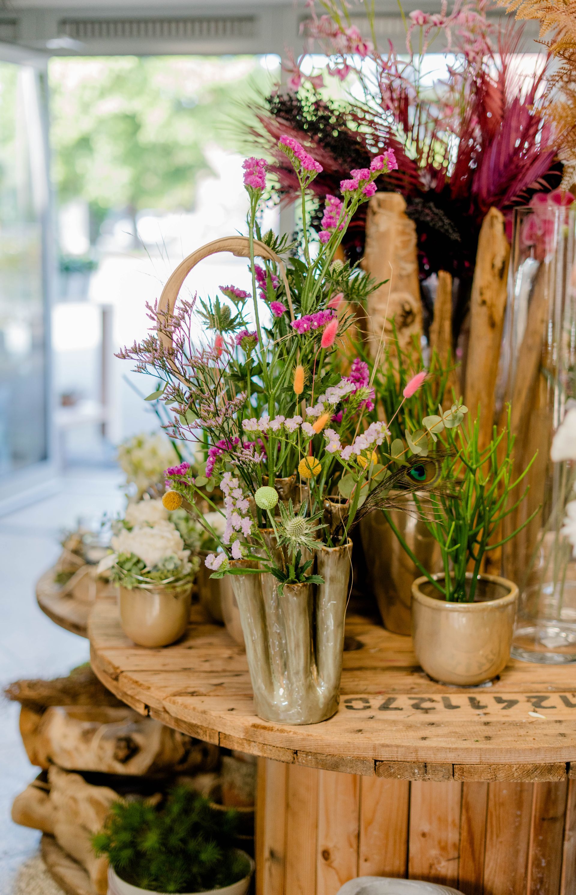 Verschiedene Blumen und Grünpflanzen sind in Vasen auf einem rustikalen Holztisch in einem hellen Laden arrangiert.