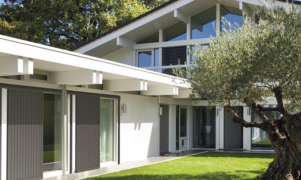 Maison blanche moderne avec volets verticaux gris foncé, fenêtres en verre et pelouse verte.