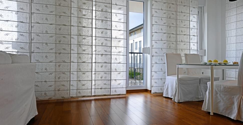 Pièce lumineuse avec stores à panneaux, porte vitrée et table/chaises blanches.