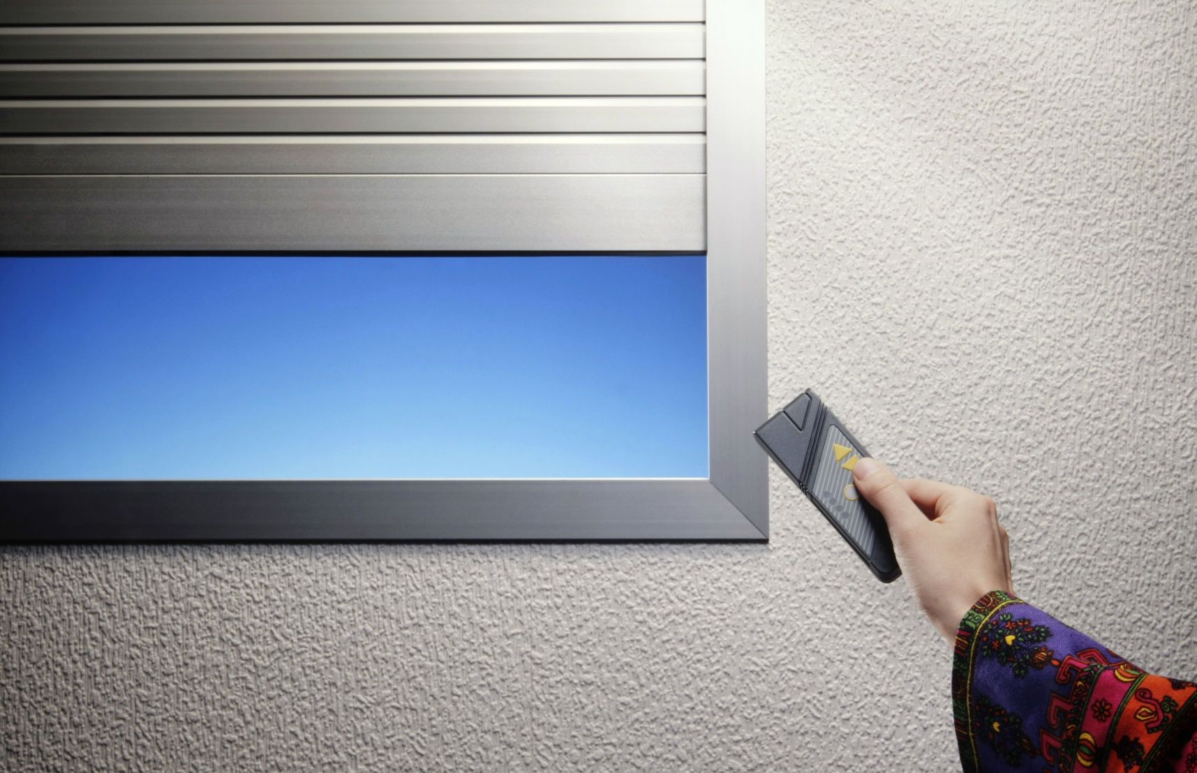Une main tenant une télécommande actionne un volet métallique surplombant un ciel bleu.