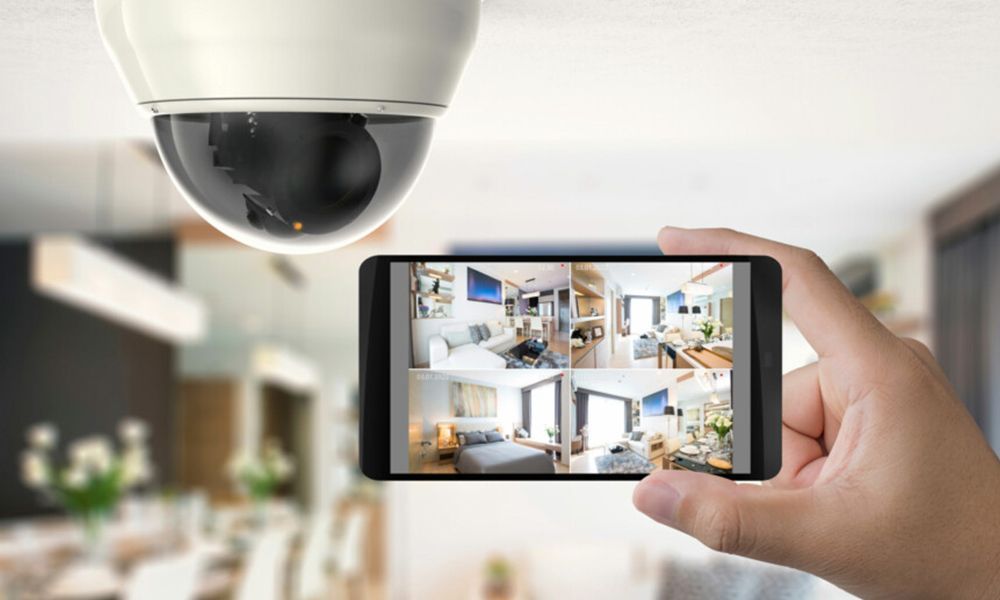 Une personne tenant un smartphone affichant les images d'une caméra de sécurité intérieure, la caméra étant visible au-dessus.
