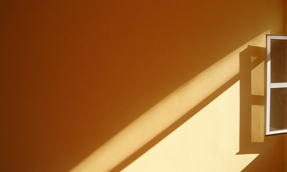 La lumière du soleil projette une ombre en diagonale sur un mur beige, avec une fenêtre entrouverte à droite.