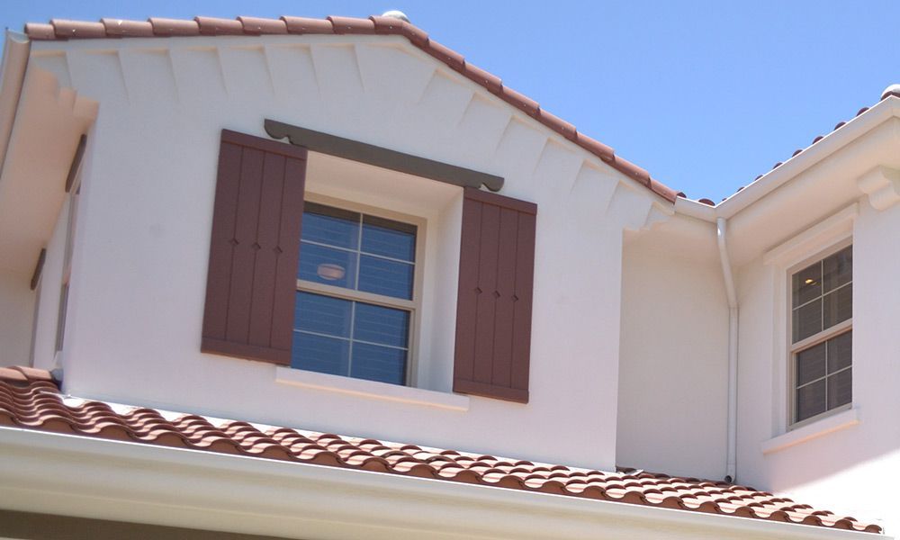 Extérieur de la maison avec toit de tuiles rouges, murs en stuc blanc, volets marron et ciel bleu clair.