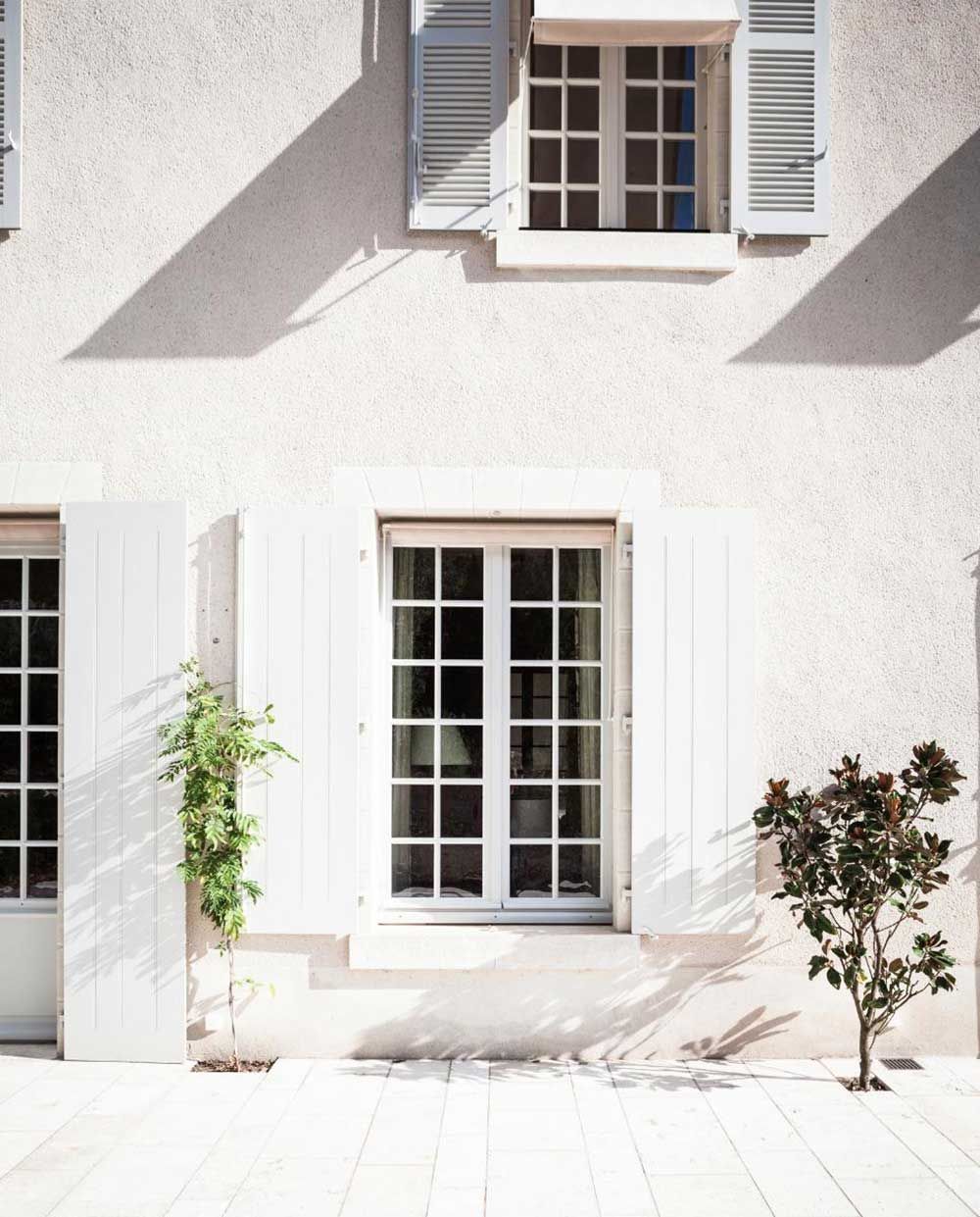 Façade d'un bâtiment blanc aux fenêtres à volets, un petit arbre et des ombres.