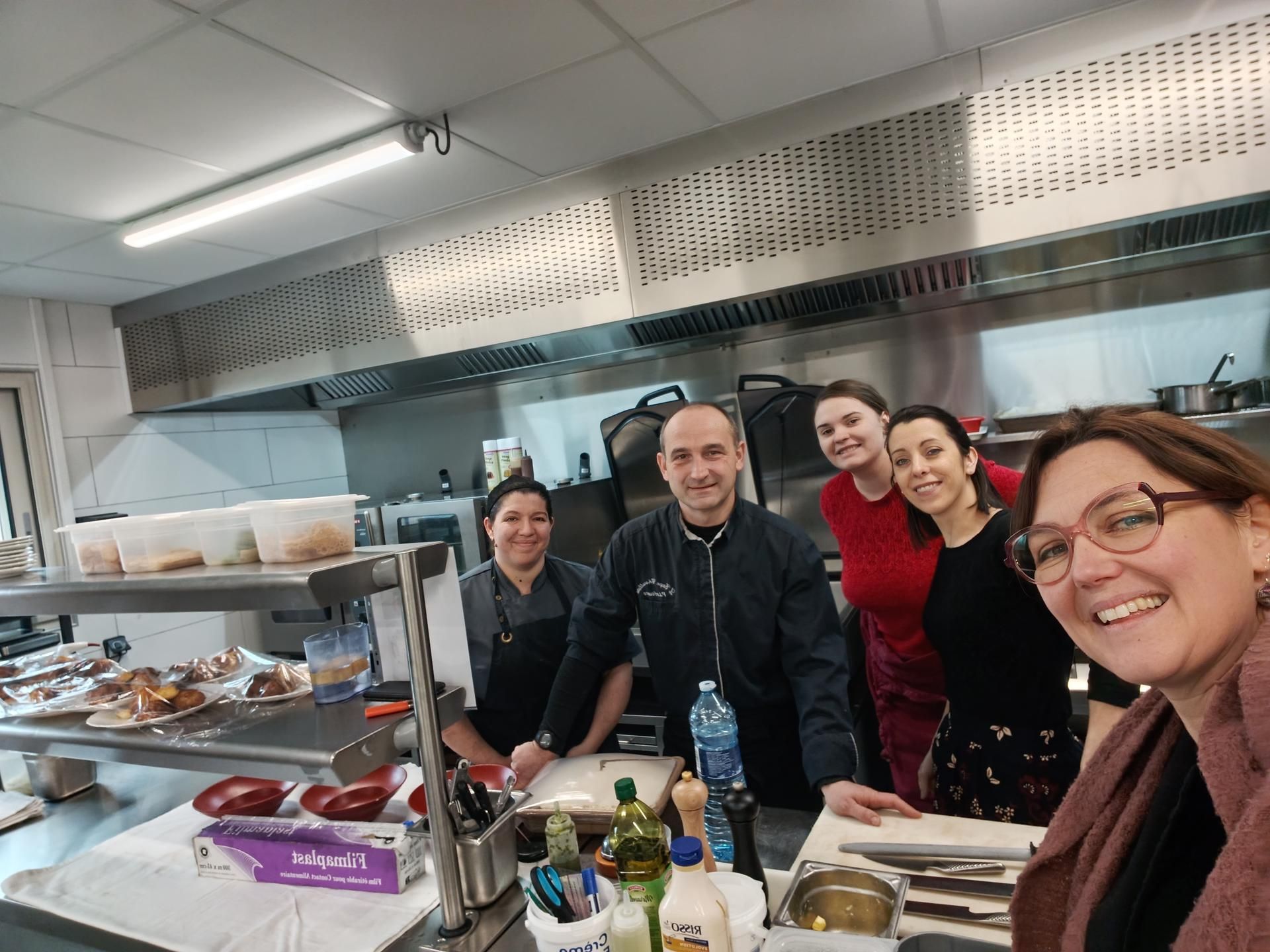 Des personnes sourient dans la cuisine d'un restaurant, équipée d'appareils en acier inoxydable et d'espaces de préparation des aliments.