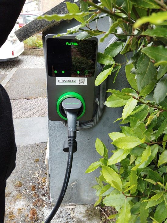 Borne de recharge pour véhicules électriques avec un câble branché à côté de buissons feuillus.