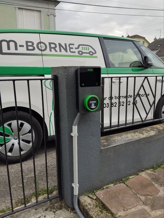 Une voiture électrique verte se recharge dans une allée, à côté d'une camionnette blanche et d'un portail noir, avec une borne de recharge murale.