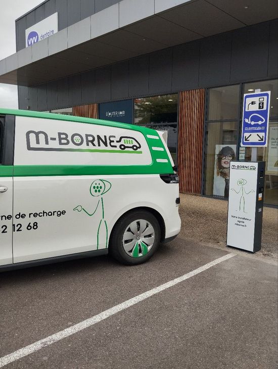 Fourgonnette électrique M-Borne stationnée à une borne de recharge à l'extérieur d'un bâtiment, avec un poteau de recharge à proximité.