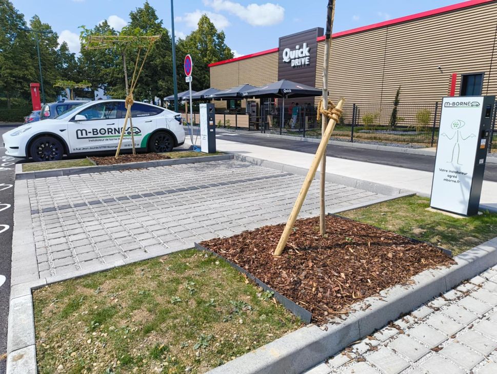 Parking devant un magasin Clarks avec une voiture blanche, un trottoir et un jeune arbre dans un parterre de paillis