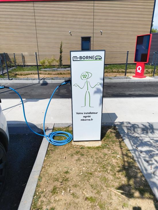 Borne de recharge extérieure pour véhicules électriques avec un chargeur sur pied blanc et un câble bleu, située en bordure de trottoir.