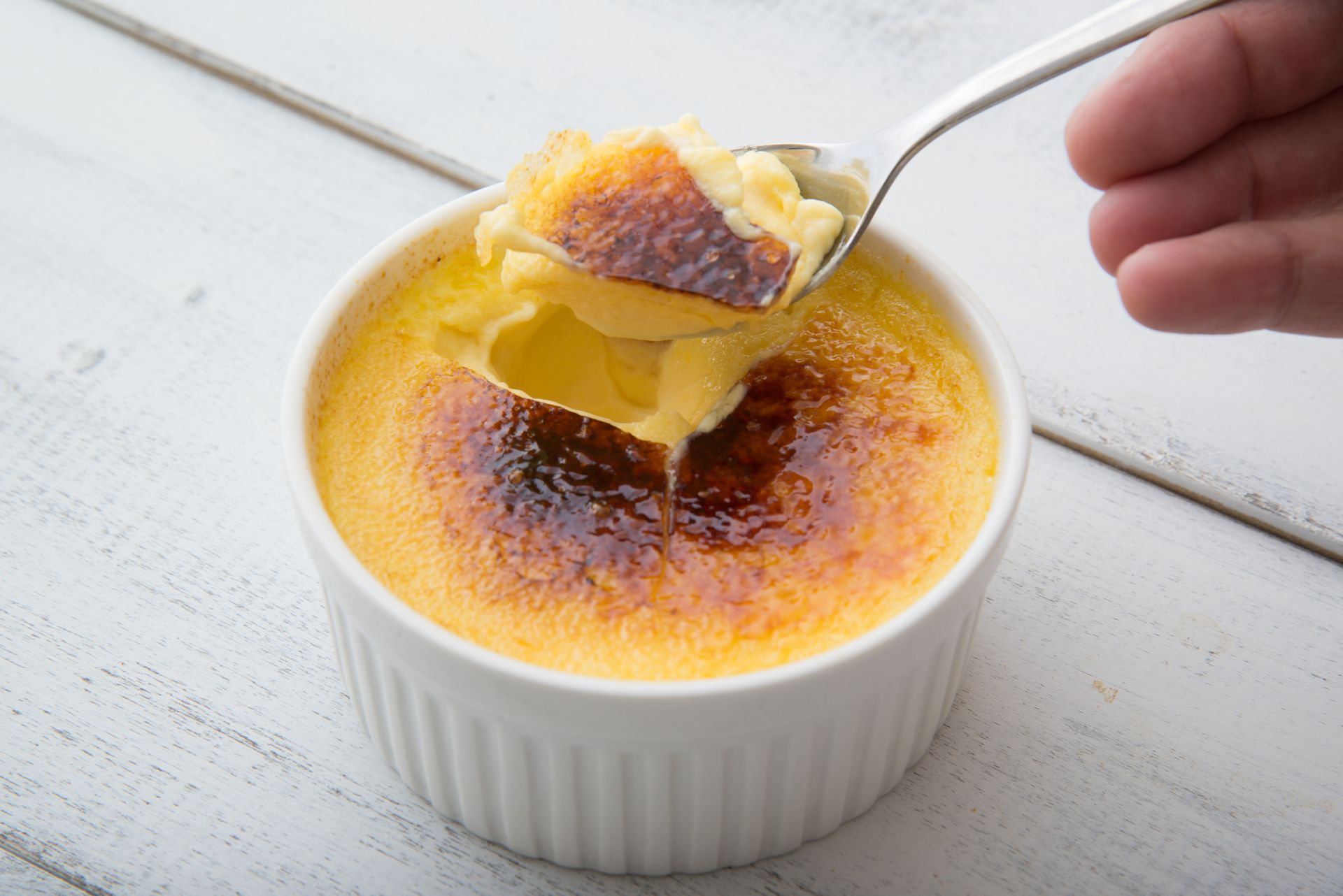 Une personne mange à la cuillère une crème brûlée caramélisée dans un ramequin blanc