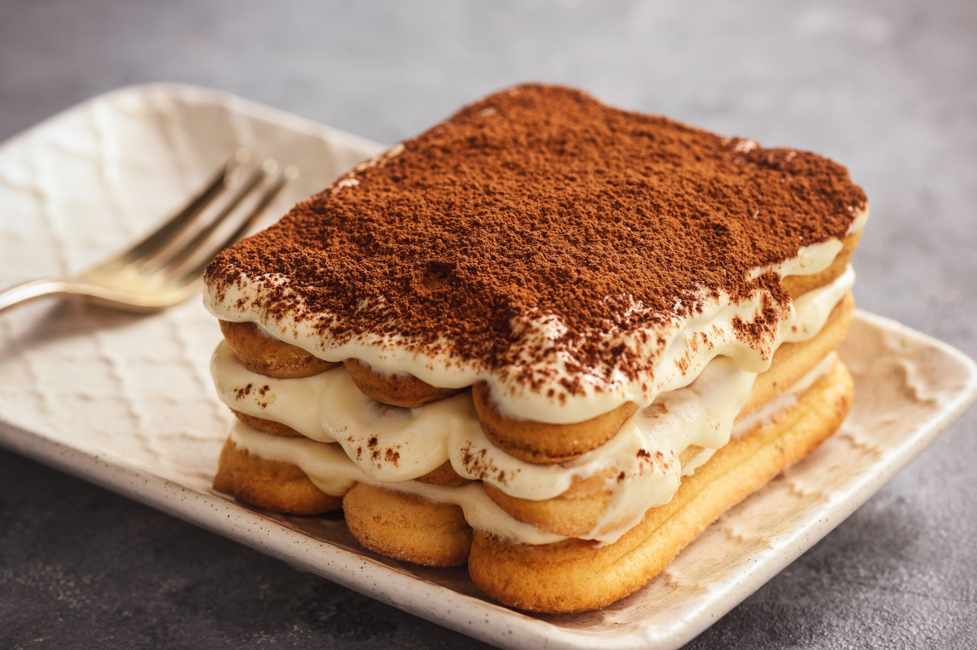 Un tiramisu servi sur une assiette carrée, saupoudré de cacao, à côté d'une fourchette sur une table grise