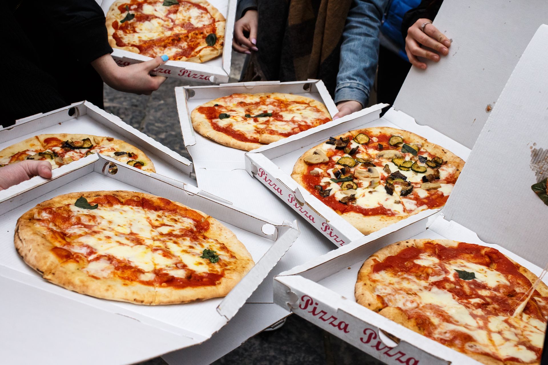 Des boîtes à pizza ouvertes contenant des pizzas assorties sont posées sur une table, au milieu de plusieurs personnes