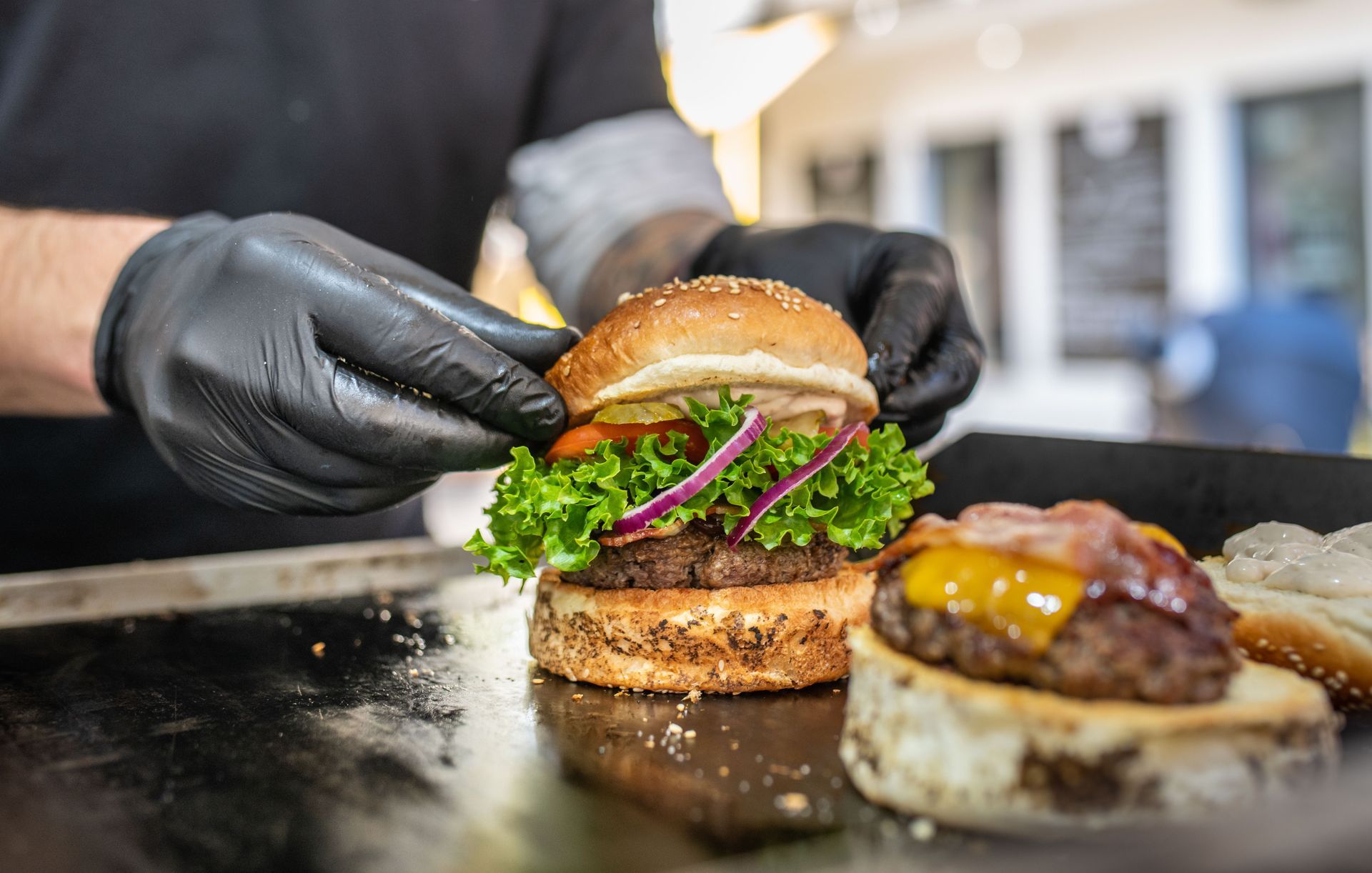 Des mains gantées assemblent un hamburger avec de la laitue et des oignons sur une plaque chauffante, un autre hamburger étant placé à côté