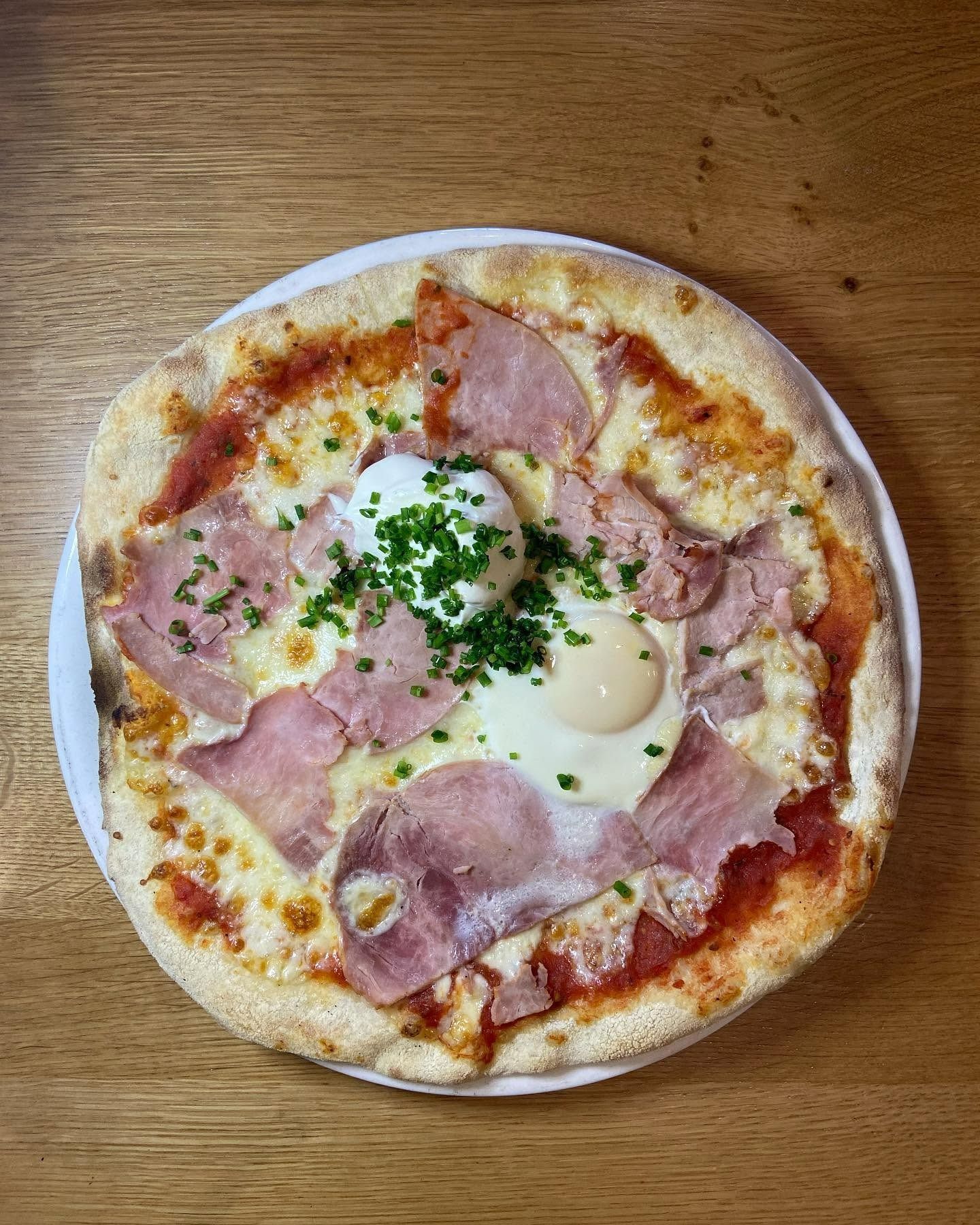 Pizza garnie de jambon, d'œuf et d'herbes sur une assiette blanche posée sur une table en bois
