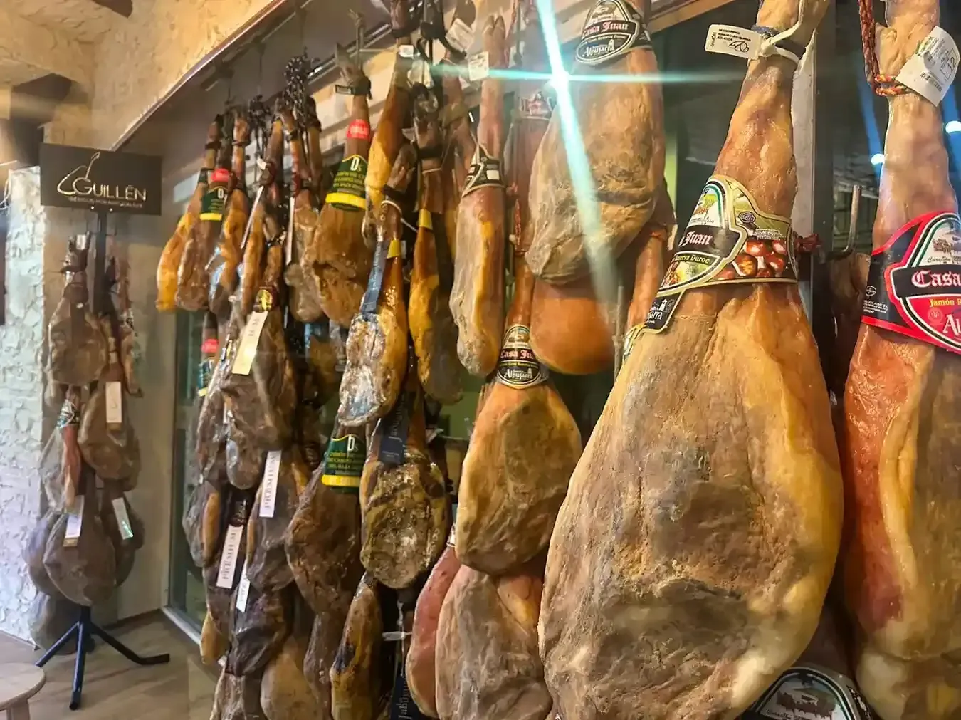Jamón curado con etiqueta roja colgando de un soporte de madera junto a una calle soleada.