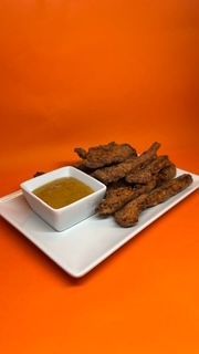 Tiras de comida frita en un plato blanco con un recipiente cuadrado de salsa amarilla para mojar, fondo naranja.