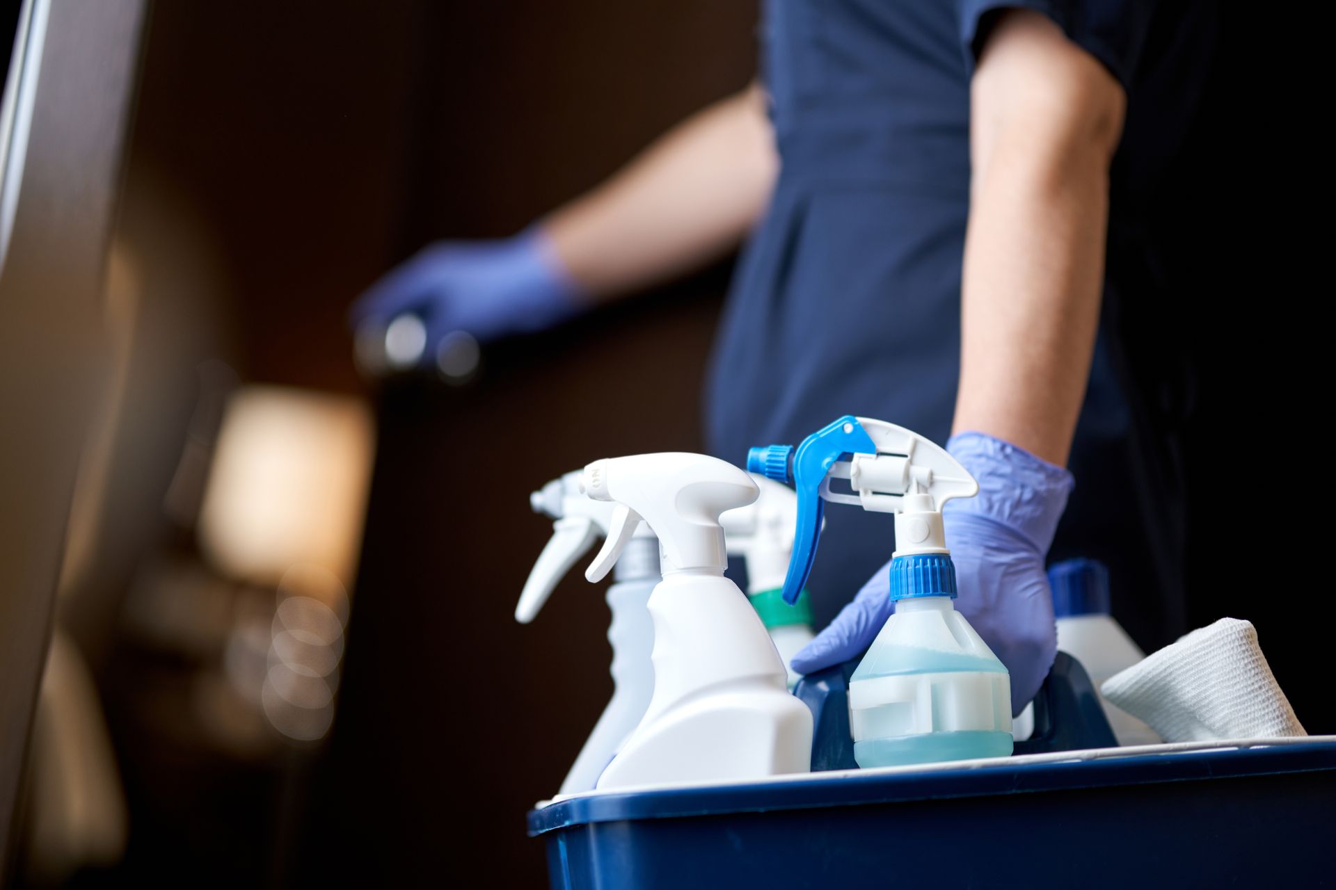 Un agent de nettoyage en uniforme sombre et gants bleus se dirige vers une porte en tenant un panier de produits de nettoyage.