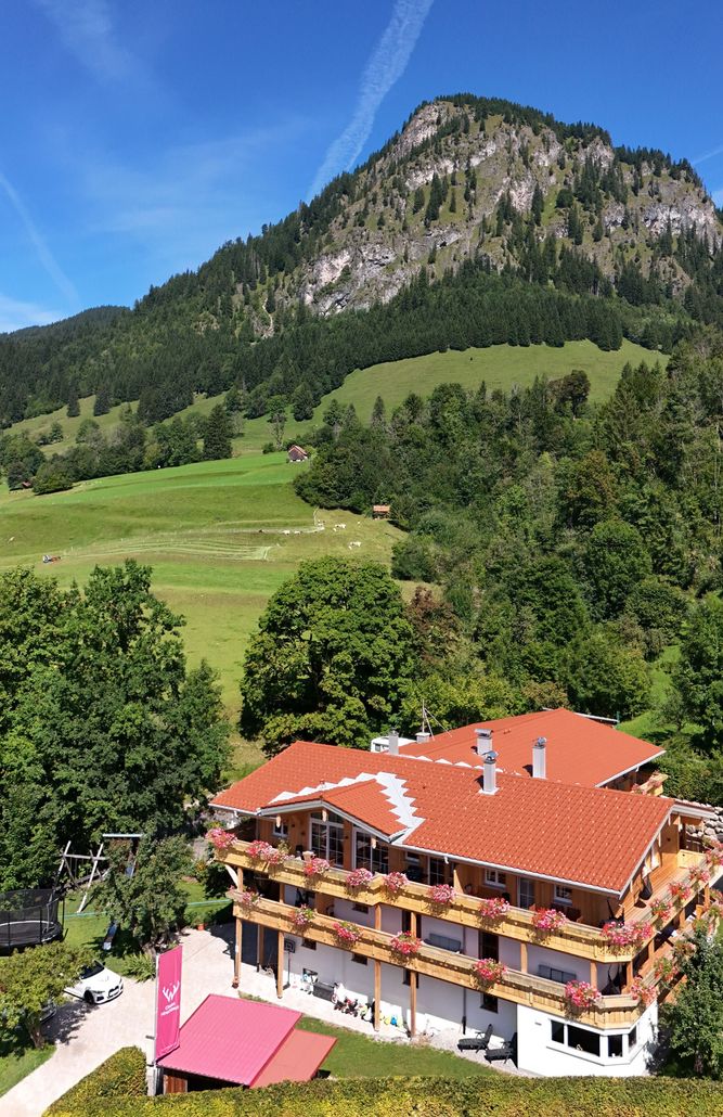 Ein großes Holzhaus ist von Bäumen, Wiesen und Bergen umgeben.
